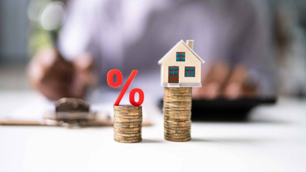 Stacked coins with a percentage symbol and a house model, illustrating the decision between remortgaging now or waiting for potential rate cuts in the UK