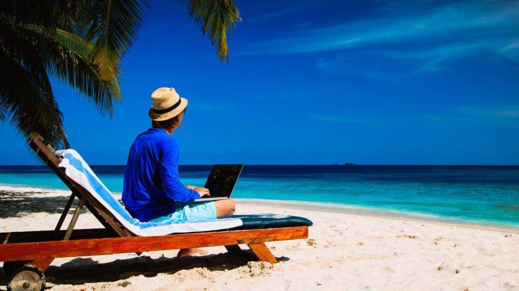 Person working remotely on a laptop at a beach, highlighting the debate on whether remote workers should be paid less