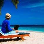 Person working remotely on a laptop at a beach, highlighting the debate on whether remote workers should be paid less