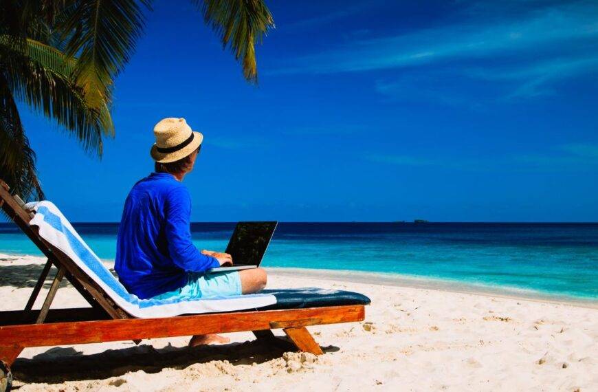 Person working remotely on a laptop at a beach, highlighting the debate on whether remote workers should be paid less