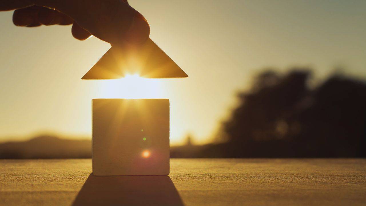 Hand placing a roof on a small house model with the sun setting in the background, symbolizing the importance of a Lady Bird Deed